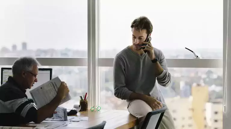 Two men in a windowed office talking; one is sitting on the desk on the phone